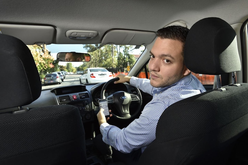 Smiling man wearing blue and white striped shirt in car looking back with one hand holding mobile phone. 