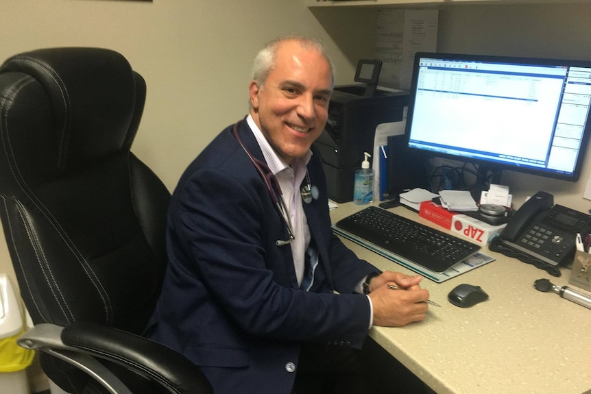 A middle-aged doctor smiles at camera while sitting at desk with computers