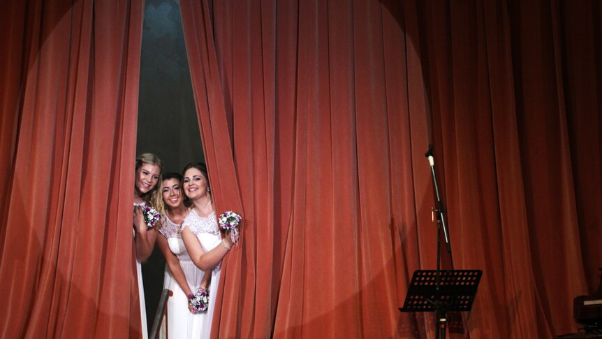 Three young women peek through heavy red velvet curtains on stage.
