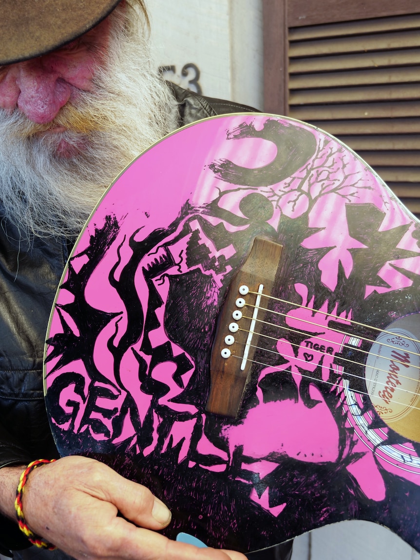 Leon Bozanich looking down away from the camera holding a pink guitar with black writing and designs