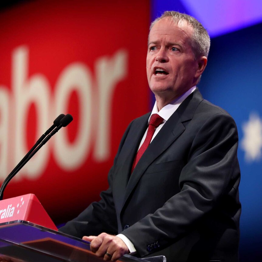 Bill Shorten stands at a lectern in front of a large sign that says Labor in red. He is wearing a red tie.