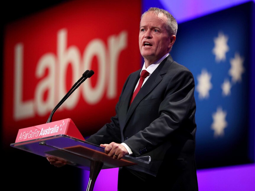 Bill Shorten stands at a lectern in front of a large sign that says Labor in red. He is wearing a red tie.