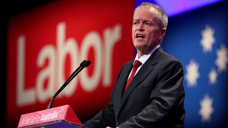 Bill Shorten stands at a lectern in front of a large sign that says Labor in red. He is wearing a red tie.