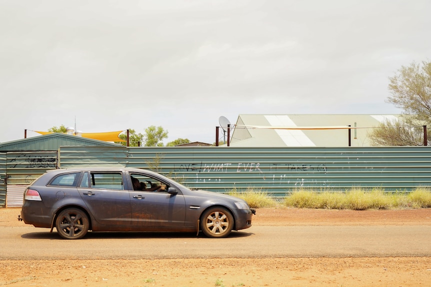 A blue Holden Commodore wagon drives in front of a graffitied fence that reads 'Love! Whatever it takes'