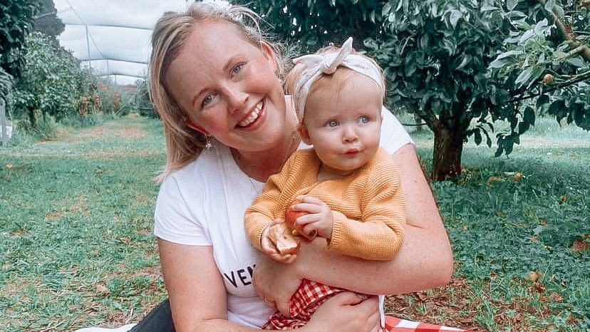 A blonde woman sits on a picnic rug smiling and holding a baby in an apple orchard.