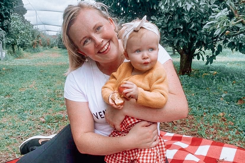 A blonde woman sits on a picnic rug smiling and holding a baby in an apple orchard.