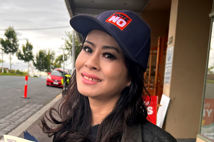 Sunny Lu smiles, wearing a 'VOTE NO' cap as she stands on a suburban street.