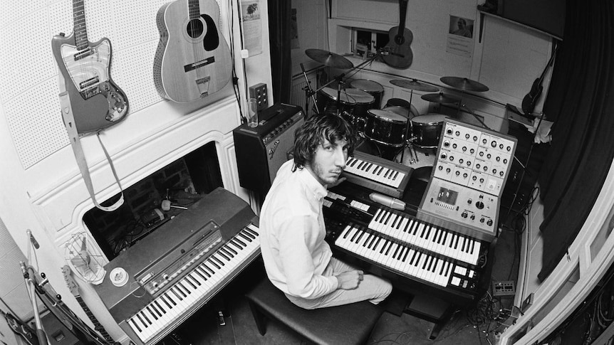 A man in a white shirt sits in the middle of a small music room surrounded by instruments like keyboards, guitars and drums.