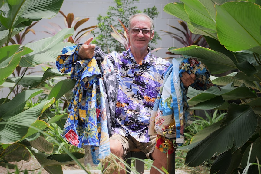 Man sitting in garden wearing tropical shirt and holding several other tropical shirts.