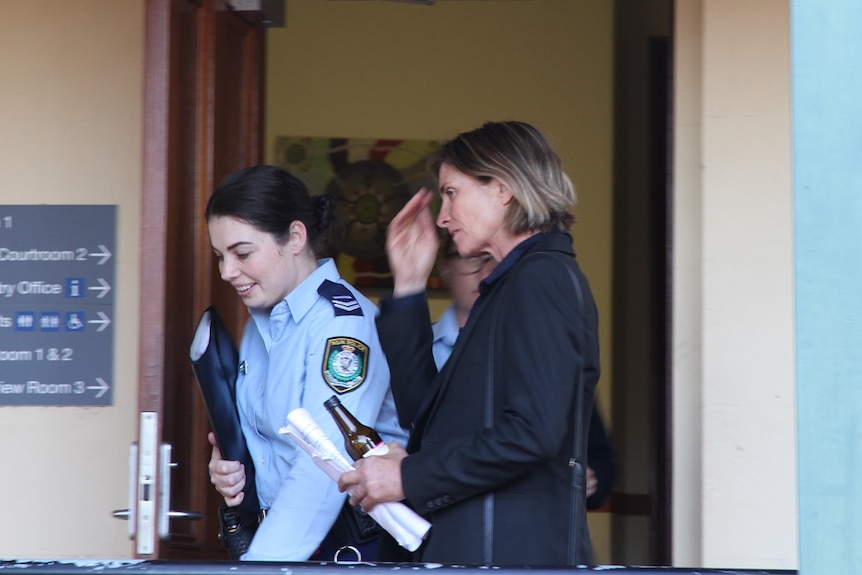 A woman with short brown hair in a black jacket walking out of court next to a female police officer.