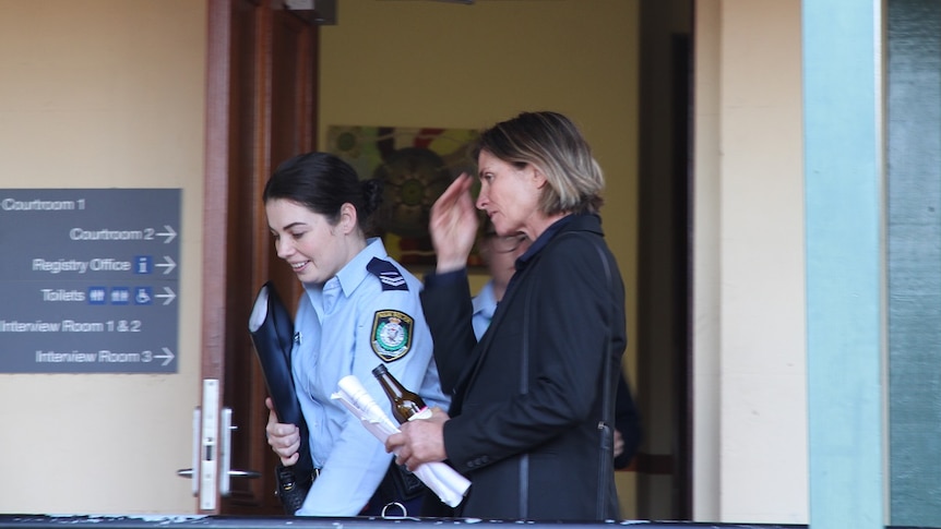 A woman with short brown hair in a black jacket walking out of court next to a female police officer.