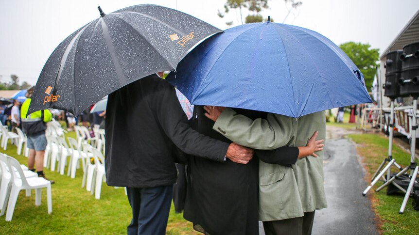 Three memorial attendees embrace under their umbrellas.