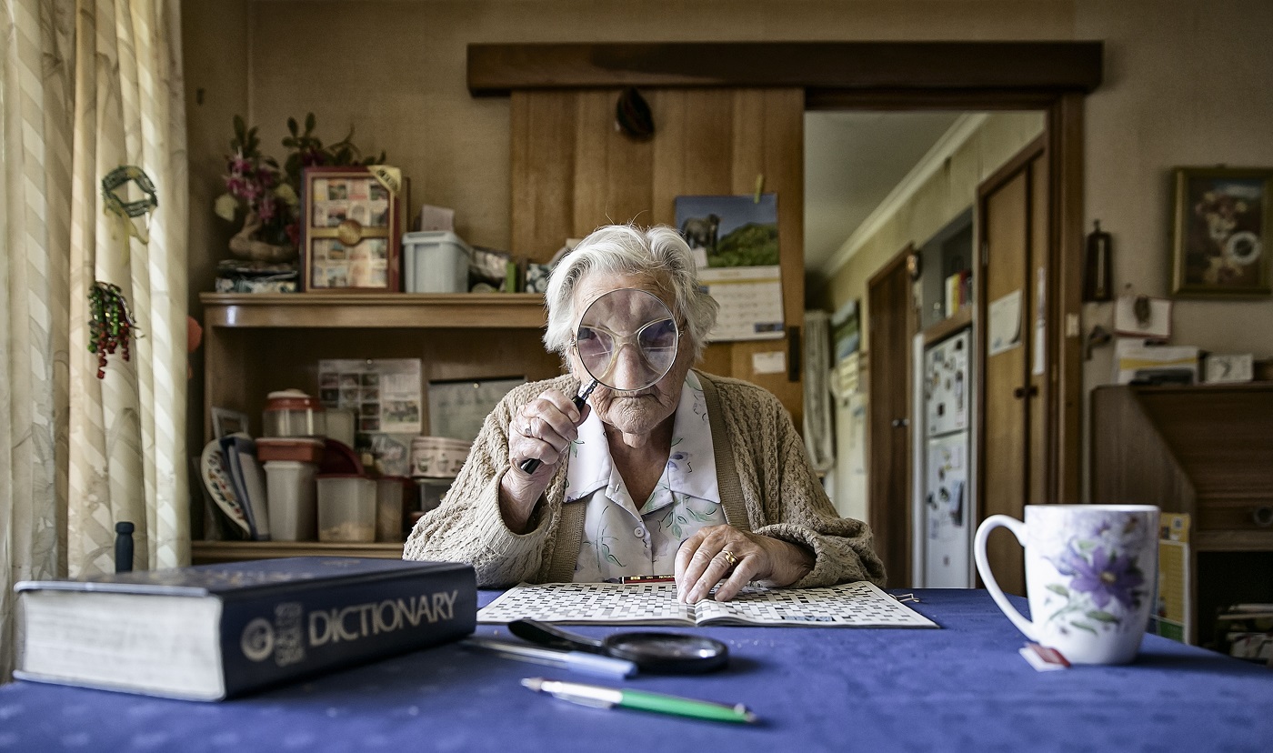An elderly woman looks at the camera through a magnifying glass, distorting her face.