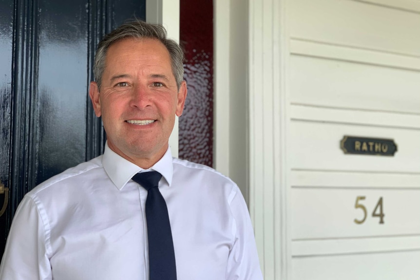 A man in white shirt and tie stands in front of a door