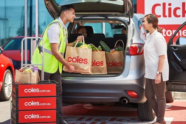 young man putting Coles shopping bags in the boot of the car, he's smiling at a woman opposite him in white shirt, brown pants