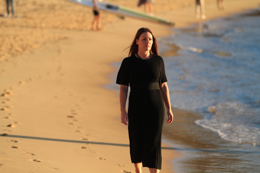 A woman wearing a black dress walks along the sand on a beach.