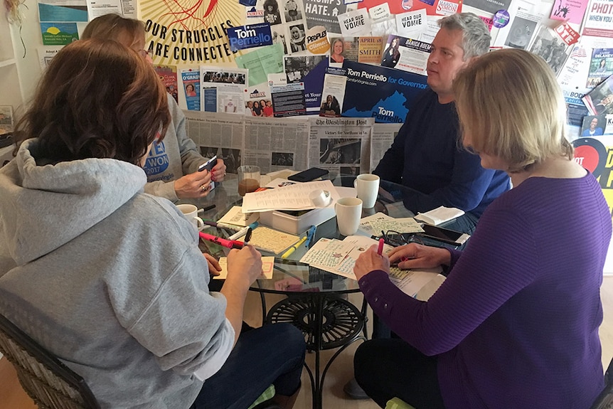 Four people sit around a table writing out postcards, with Democratic promotional material on the wall behind them.