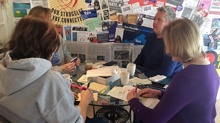 Four people sit around a table writing out postcards, with Democratic promotional material on the wall behind them.