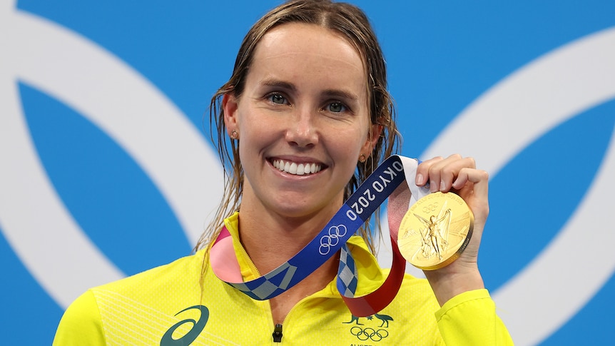 Emma McKeon smiles while holding up her gold medal. She's wearing her gold Australian jacket