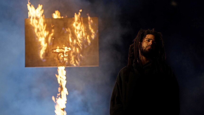 Photograph of J. Cole with basketball ring in background on fire.