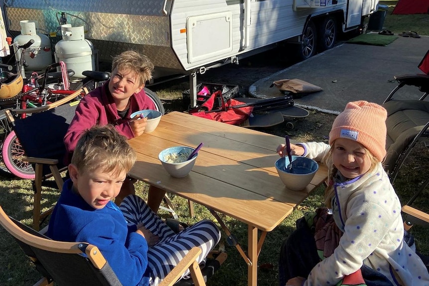 Three children sitting at outdoor table in front of a caravan.