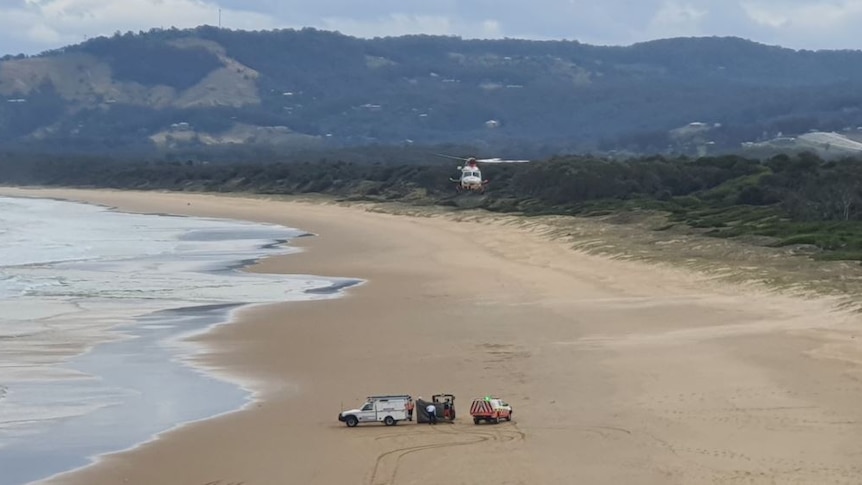 A helicopter flies above emergence vehicles parked on a beach.