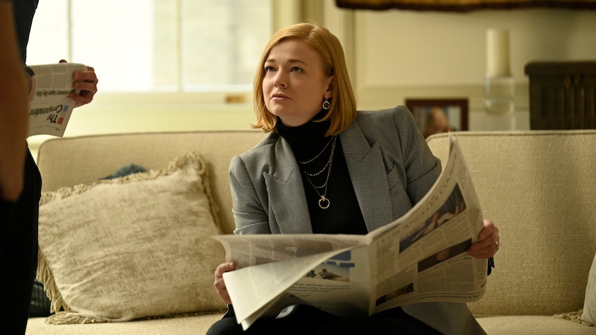 Sarah's character Shiv sits on a couch and looks up at someone off camera while holding a newspaper.