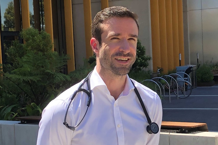 a man with a stethoscope over his shoulders and a white shirt stands outside a building