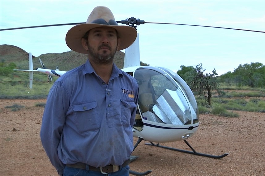 A man in a blue shirt and a cowboy hat stands near a helicopter