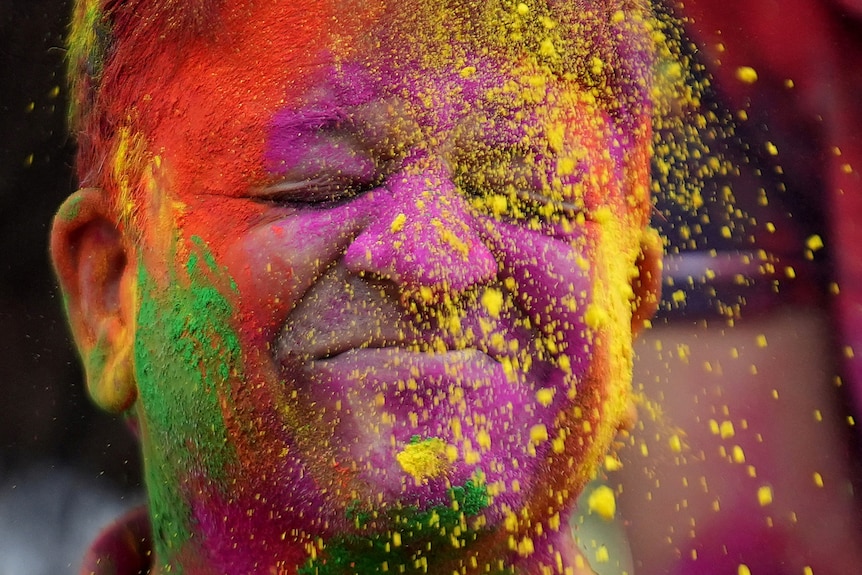 A man closes his eyes as pink, green and yellow powder is thrown on his face as part of a ritual celebration.