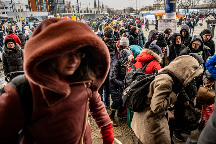 A long line of people, stand five abreast in thick winter coats.  