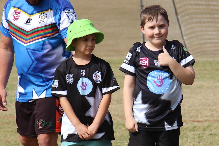 Two young boys in sport uniforms, one giving a thumbs up, with a male coach in the background