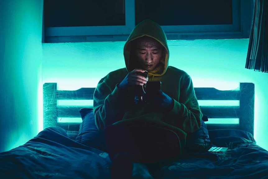 Man in hoodie looking at phone while sitting on bed for a story about what to do when you're being stalked.