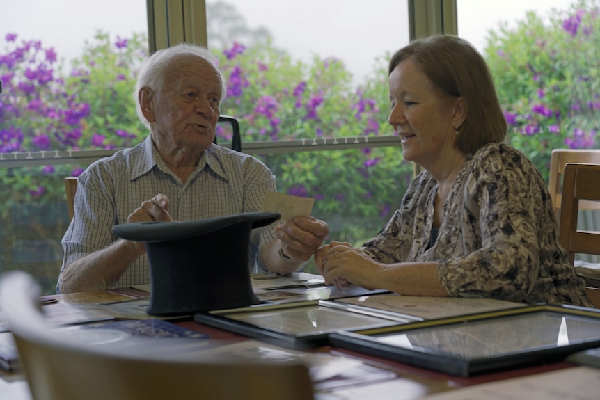 An elderly man holds up a photograph and smiles while sitting at kitchen table