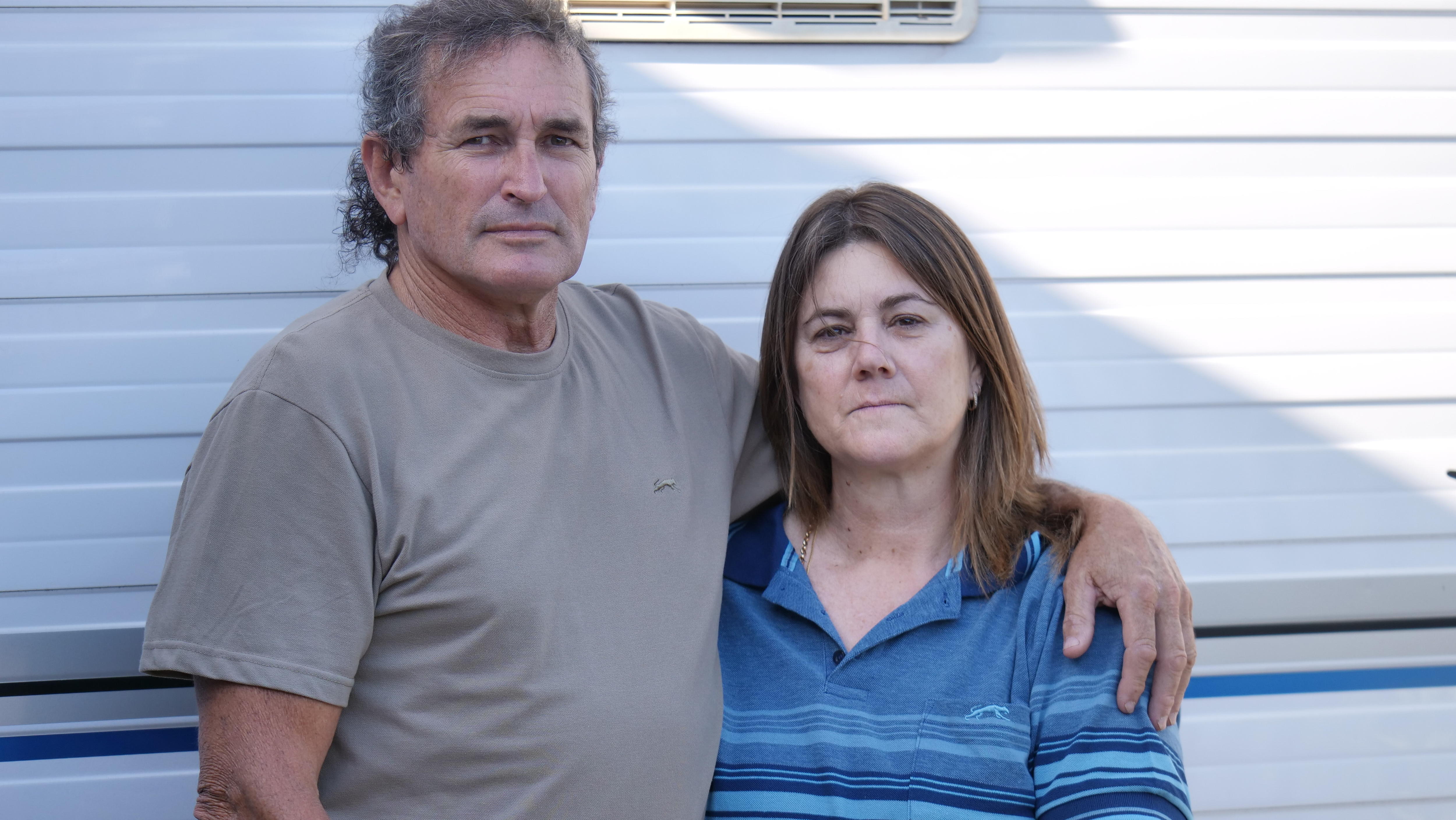 Un hombre se encuentra frente a una caravana con su brazo alrededor de una mujer, ambos mirando sombríamente a la cámara.