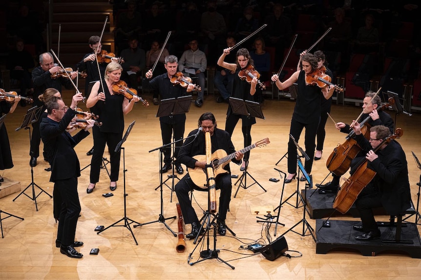 a chamber orchestra performs surrounding a didgeridoo player on a stage