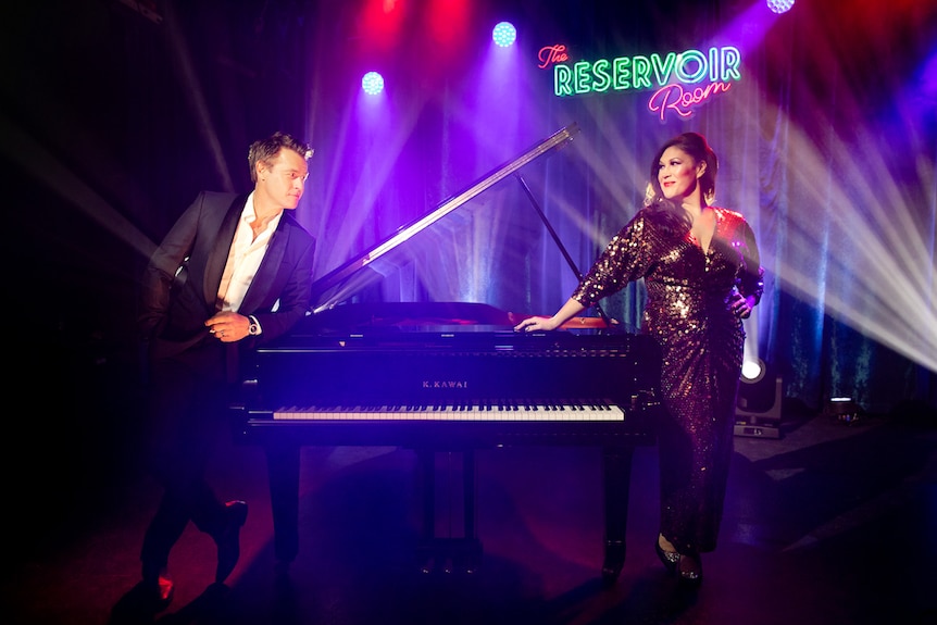 Darkened studio lit with purple and red spotlights, piano in centre with man and woman leaning either side.