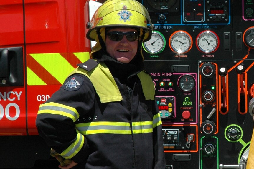 A firefighter in uniform and helmet stands in front of a fire truck wearing sunglasses