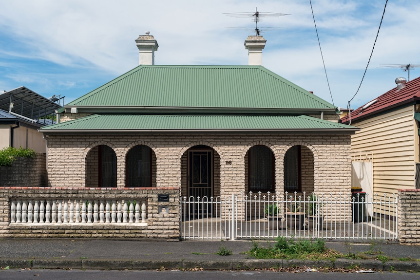 You view a brown brick veneer house with two white chimneys and green, corrugated iron roof on a clear day.