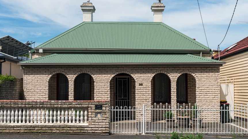 You view a brown brick veneer house with two white chimneys and green, corrugated iron roof on a clear day.