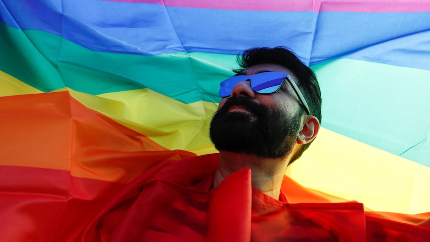 A bearded man wearing sunglasses holds a rainbow LGBT flag aloft.