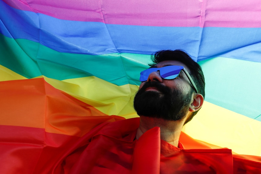 A bearded man wearing sunglasses holds a rainbow LGBT flag aloft.