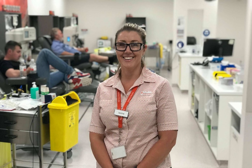 A woman stands smiling in the middle of a blood bank while donors recline behind her in large chairs.