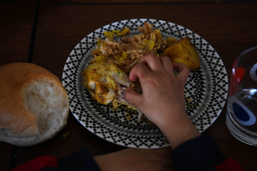 Close up of a child's hands picking up a piece of chicken off a dinner plate. A half-eaten bread roll on the left