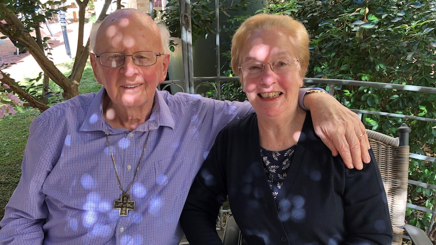 an older couple sit in the shade in a park, his arm around her shoulders