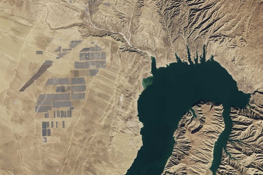 You view a satellite image of a large dam in the middle of light brown land with a large solar park visible to its left.