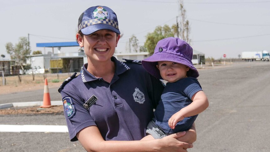 A female police officer stands in an empty street in rural Queensland holding her young son.