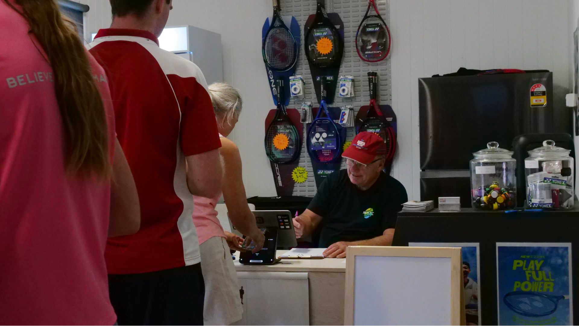 La gente hace fila para ver a Paul en el mostrador donde tiene que registrarse y pagar, con raquetas de tenis en la pared detrás de él.