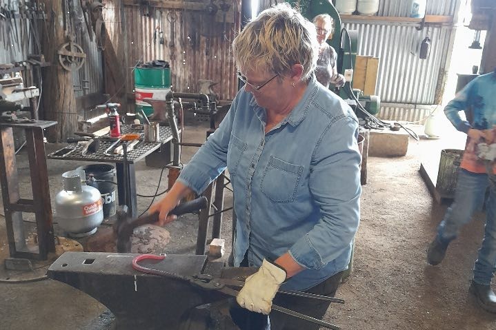 Woman in blue hammering a red hot hook on an anvil in a blacksmiths workshop 