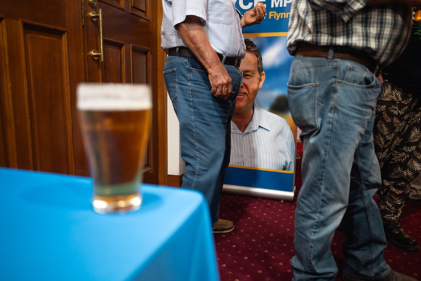 A beer sits on a table indoors. A couple of men can be seen standing nearby, one is in front of a sign for Flynn MP Colin Boyce.